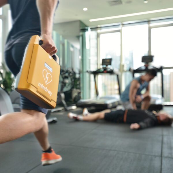 Person holding a yellow AED (Automated External Defibrillator) in a gym setting with blurred background